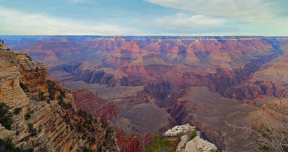 Panoramic view across the Grand Canyon at sunset, with layered pink and red rock formations beneath a pale blue sky.